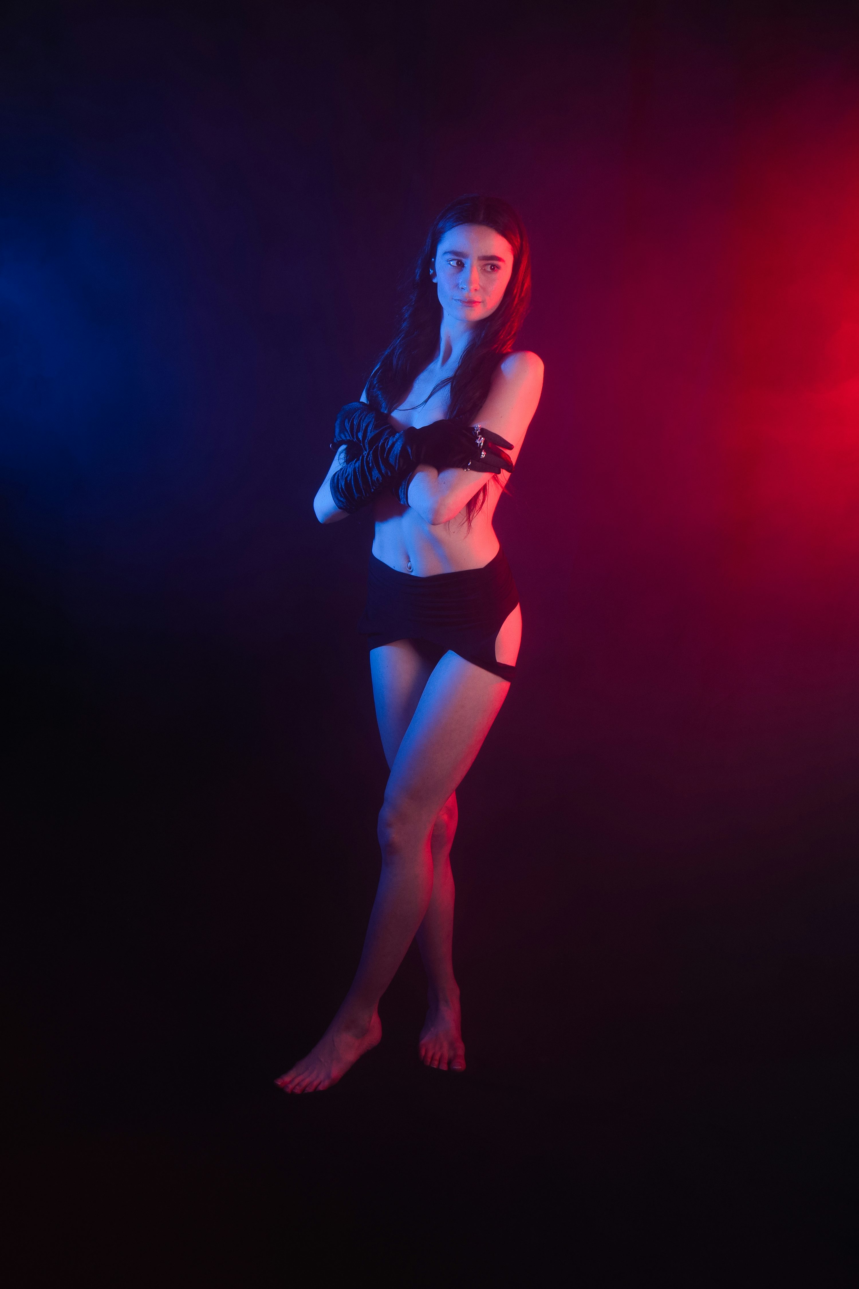 young woman stands in a dark studio against a background of red and blue neon lighting.  she is wearing a black short outfit and black gloves, her arms crossed over her chest.  her expression is confident and slightly seductive.  there is a smoky effect in the background, adding to the mysterious atmosphere.  the lighting dramatically illuminates her body and accentuates her pose.