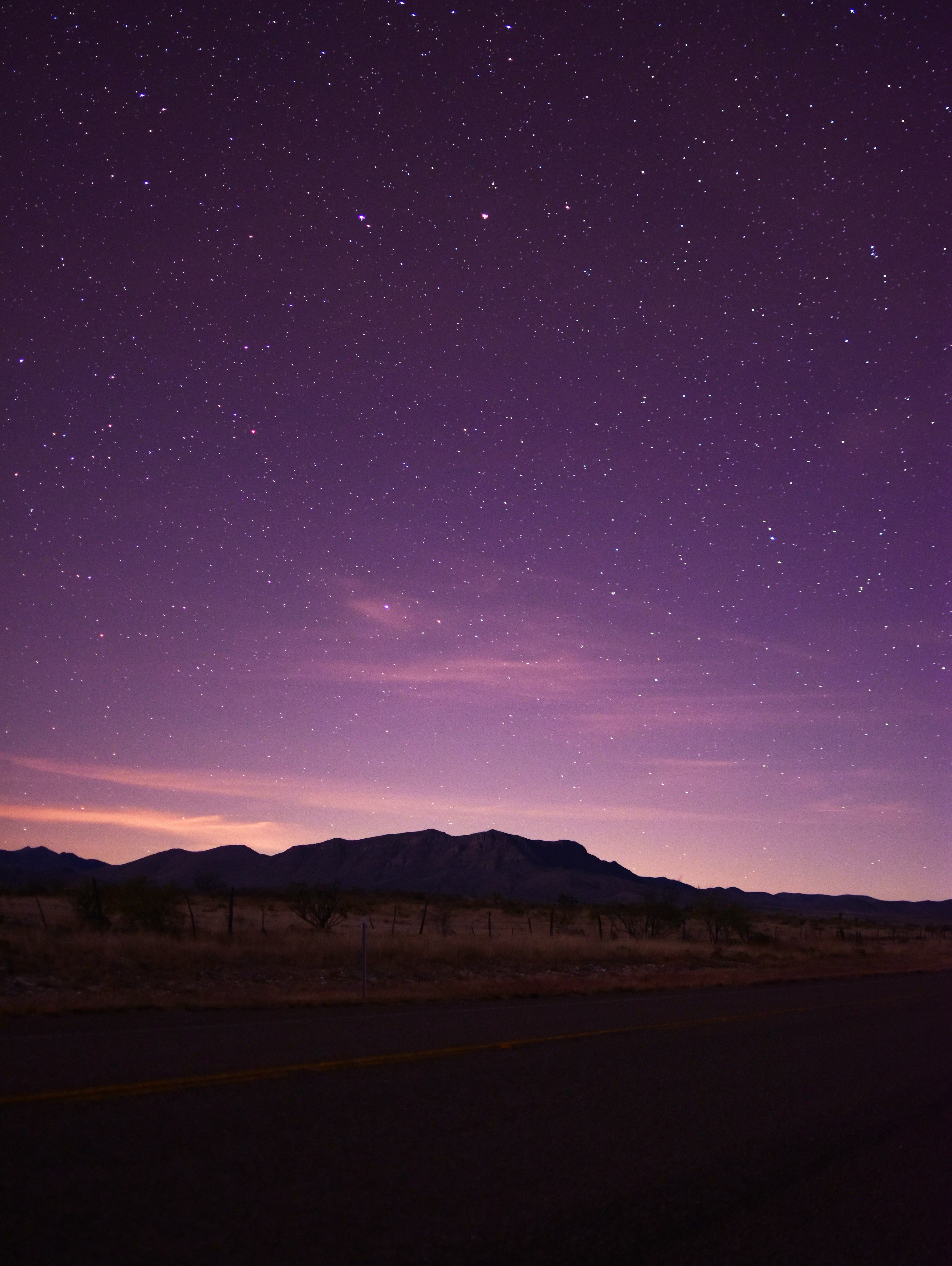 a purplehued night sky is filled with a multitude of stars. the faint outline of a mountain range sits on the horizon against the fading light of dusk. the foreground features a dark, empty road with a fence line and sparse vegetation running alongside.