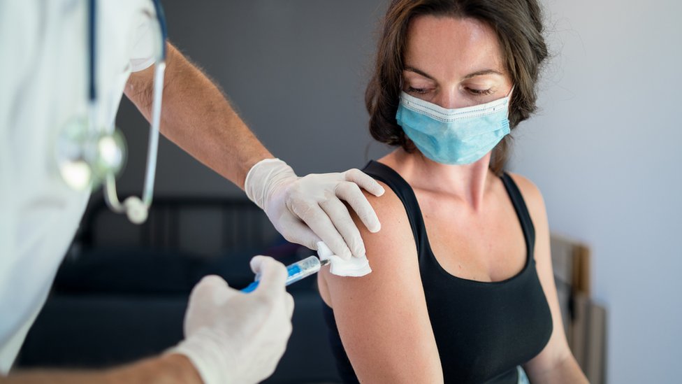 a healthcare worker is administering a vaccine to a patient. the patient is wearing a face mask and a black tank top. the doctor is wearing gloves and a white coat with a stethoscope. the vaccine is being injected into the patient's arm after being cleaned with a cotton swab.