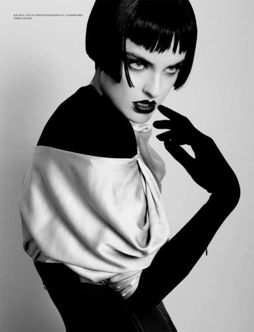 a black and white portrait of a woman with short black hair and bangs, wearing a dark outfit and gloves. she is posing with a serious expression and looking directly at the camera. the lighting is dramatic, creating strong shadows and highlighting her features.