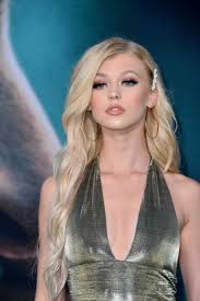 a photo of loren gray in a shimmering gold halter top. she is blonde, with her hair parted on the side. the background is a blue color.