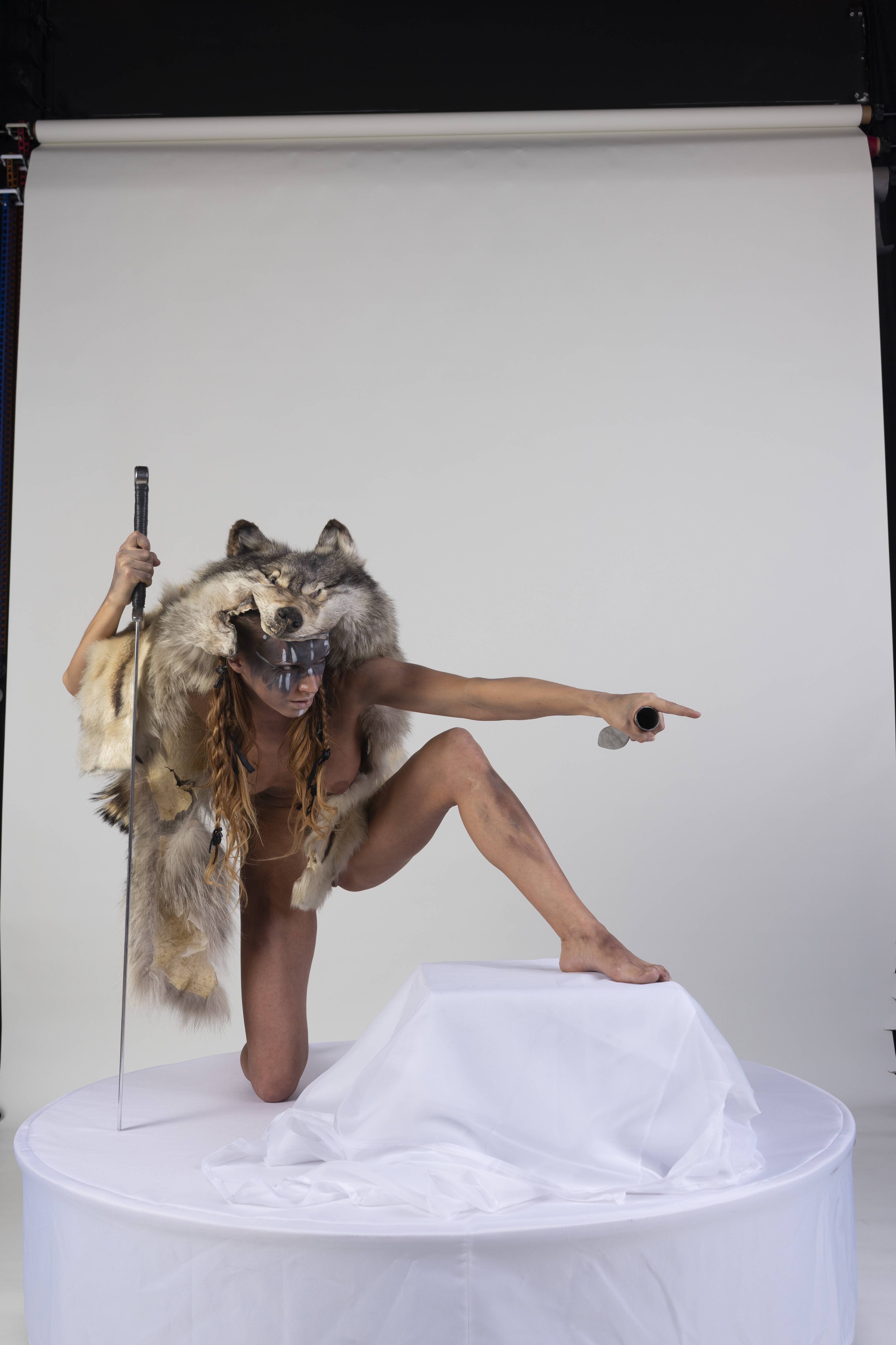 a studio shot depicts a woman in a fantasyinspired pose. she is kneeling on one knee on a white platform covered with draped white fabric, her bare torso adorned with a wolf headdress made of fur. in one hand, she holds a sword upright, while the other is outstretched, pointing towards the right. the background is a plain white studio backdrop.