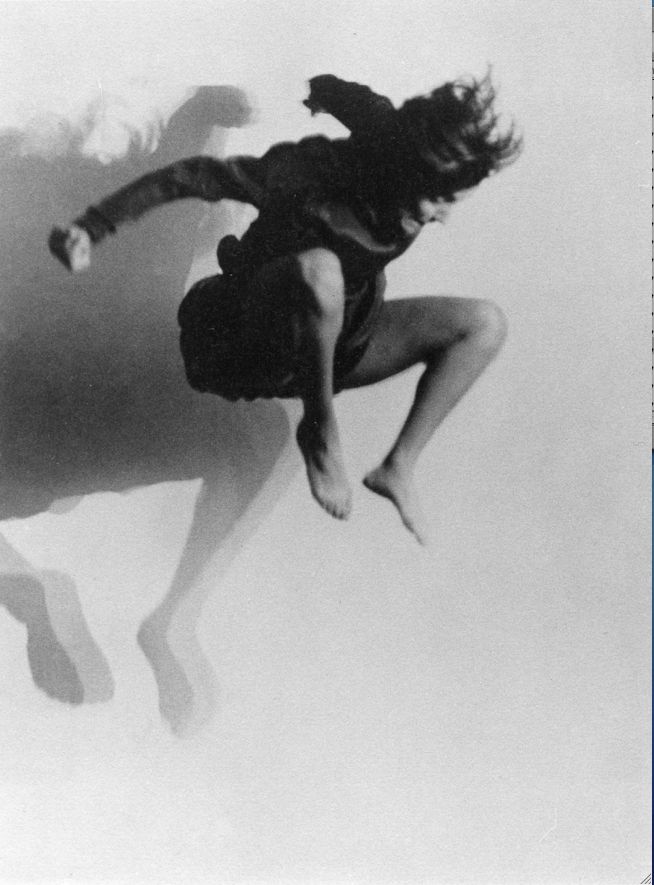 a black and white photograph of a woman mid leap, with her arms outstretched and her hair flowing behind her. she appears to be dancing,  the image is blurred and has a dreamlike quality, suggesting movement and energy. the image is framed against a white background.