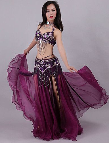 a woman wearing a purple belly dancing costume poses against a white background. she is wearing a sequined bra top, a matching skirt, and a long purple chiffon scarf draped around her waist. she is also wearing a necklace and bracelets. she is looking at the camera with a serious expression.