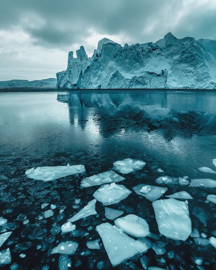 the image shows a large iceberg reflected in the water with smaller ice fragments floating in the foreground. the sky is overcast with clouds. the scene is a cold, frozen arctic landscape.
