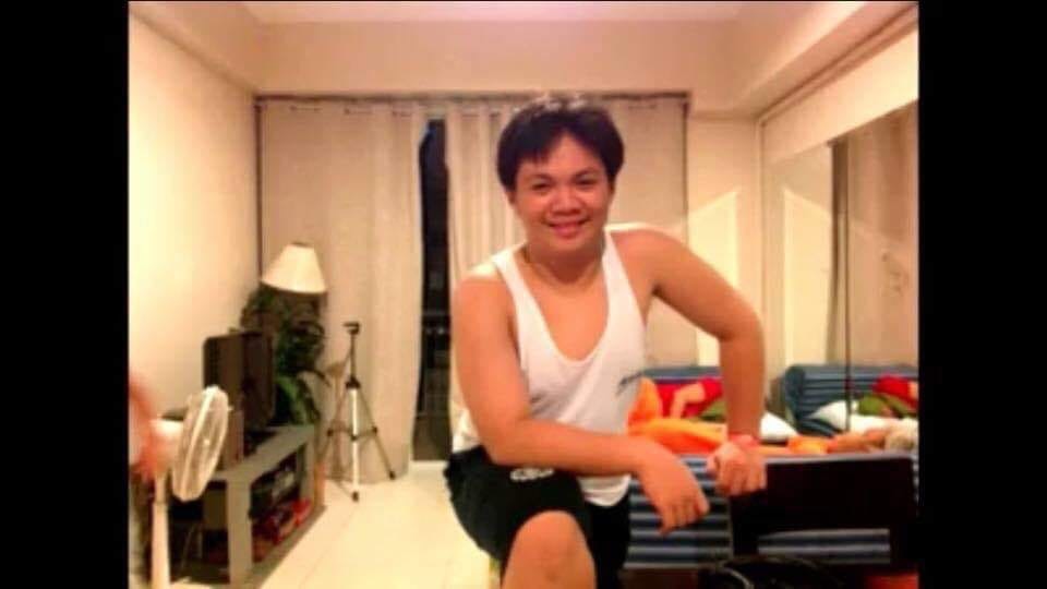 a young man is posing for a selfie in his apartment living room. he is wearing a white tank top and black shorts. the room is furnished with a couch, a coffee table, and a television. the lighting is artificial and the perspective is eyelevel.