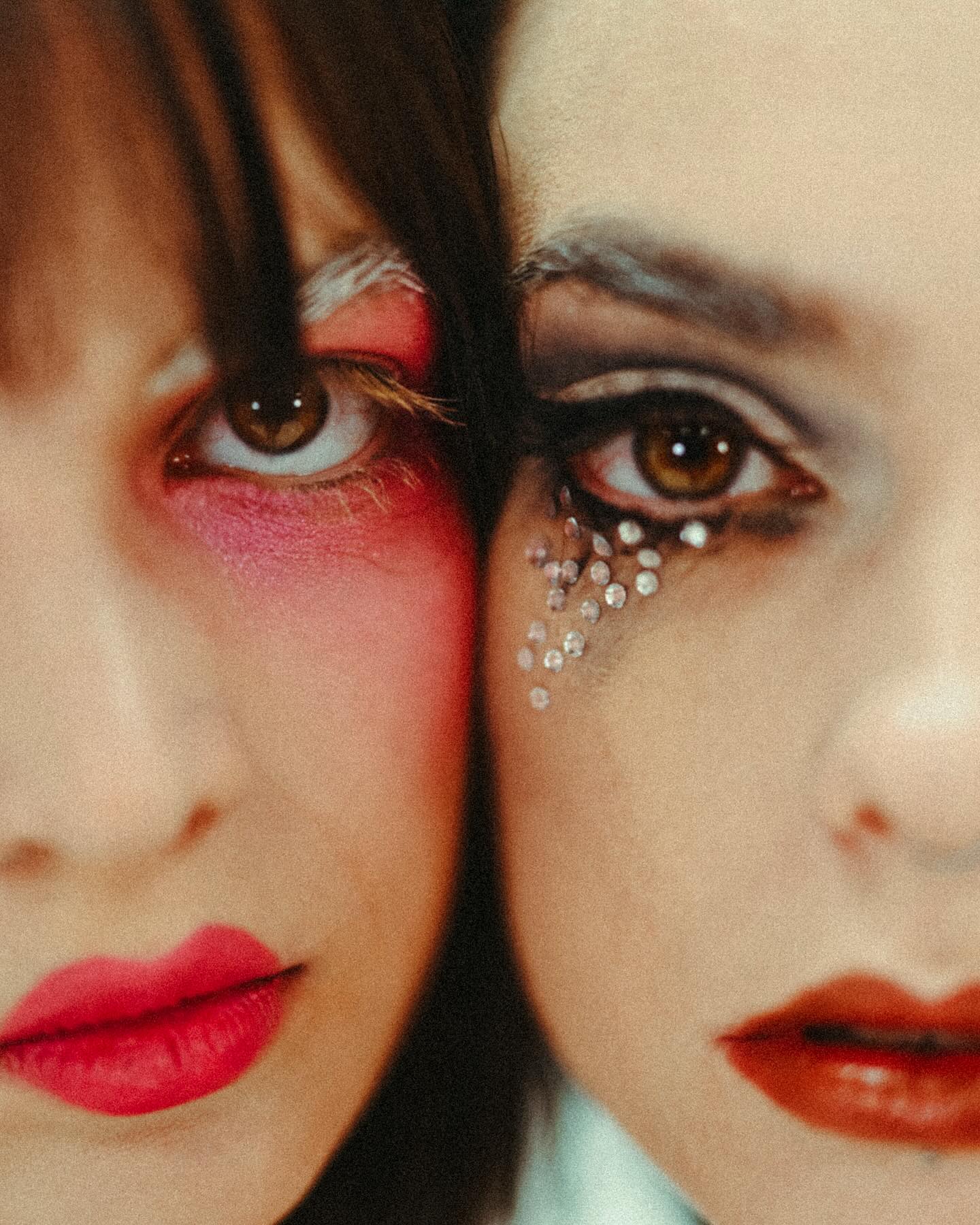 the image is a closeup shot of two women's faces pressed together.  both women are wearing striking and contrasting makeup. one woman has pink and red makeup on her face, including bright red lips.  the other woman features dark eyeshadow, glitter, and orangetoned lipstick. the makeup is theatrical and artistic in style. the focus is sharply on their faces, highlighting the texture of their skin and the details of their makeup.  the overall mood is intimate and visually striking.