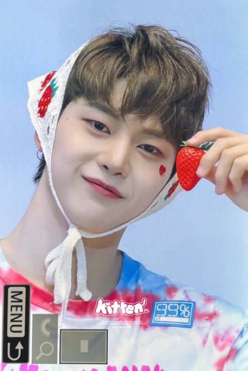 a closeup selfie of a young man with brown hair wearing a white crocheted strawberry hat. he is holding a strawberry near his face and smiling softly at the camera. he has a small red heart drawn on his cheek. he is wearing a colorful tshirt with a "kitten" logo and japanese writing. the background is a pale blue. the photo gives off a soft and playful vibe.