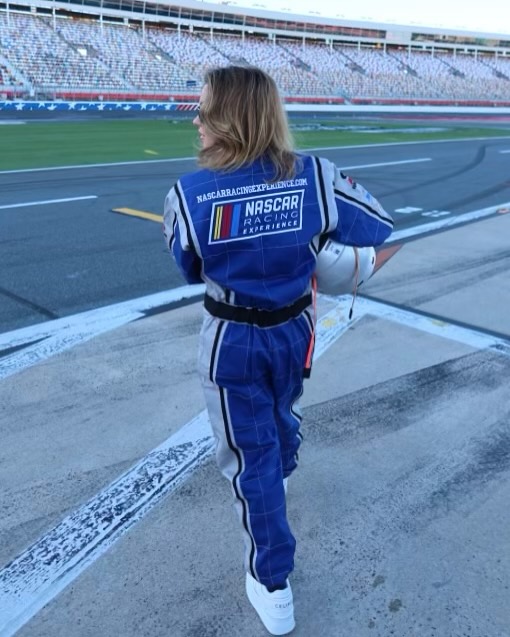 the image shows a woman from behind, wearing a blue and beige nascar racing experience jumpsuit. she's standing on a race track with a large, mostly empty grandstand visible in the background.  she appears to be holding a helmet. the overall setting suggests she's at a nascar event or experience. the woman is facing away from the camera, and her expression is not visible. the focus is on the racing attire and the setting.
