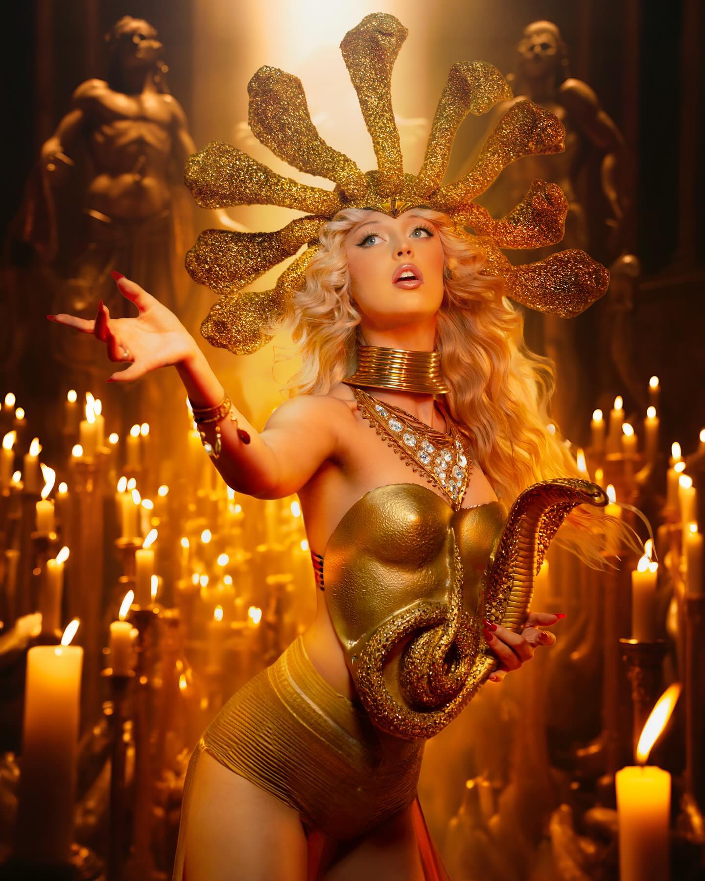 a woman with blonde hair and a golden snake headdress stands among numerous lit candles. she wears a gold costume, adorned with snakethemed jewelry and holds a decorative gold snake. two statues are visible in the background. the lighting creates a warm and dramatic atmosphere.