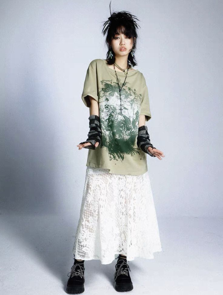 a young woman with short dark hair styled in a slightly messy fashion is posing against a plain light grey background.  she is wearing an oversized light olive green tshirt featuring a dark print, a long white lace skirt, black chunky boots and fingerless gloves. she also wears a delicate silver necklace. her pose is slightly casual, with her hands slightly out to the sides. the overall style is described as edgy and alternative.