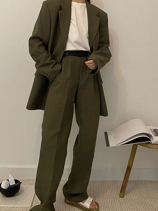a woman is posing in a stylish and modern outfit. she is wearing green pants with a matching blazer and a white shirt. she has a black belt cinched around her waist,  and white sandals on her feet. the woman looks stylish and put together. she is standing in a room with a light wood table with a book on it.