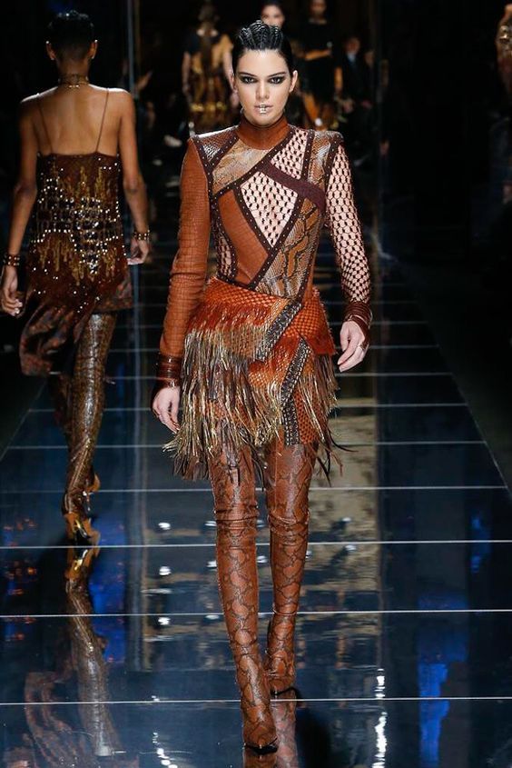 a fashion model walks the runway wearing a brown leather dress with fringe and thigh high boots. the dress is a patchwork of different leather textures and colors, and the boots are snake print. the model is accessorized with gold jewelry.