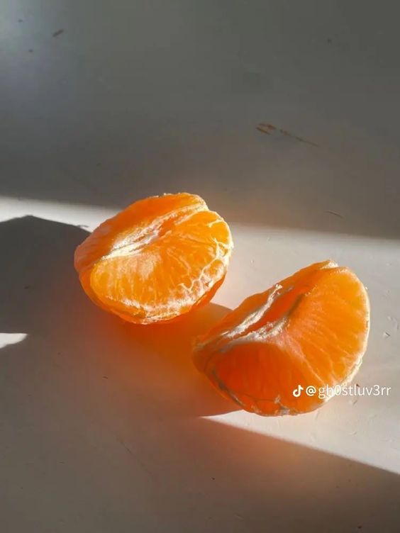 two halves of a peeled tangerine are bathed in natural sunlight, casting long shadows on a white surface. the fruit's vibrant orange segments glisten, showcasing its juicy texture.