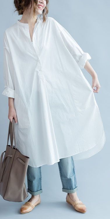 a young woman in a white shirt dress, blue jeans, and brown shoes. she's holding a brown tote bag and smiling. she's standing against a light blue background. she's looking at the camera. she's dressed in a simple and casual yet elegant way.