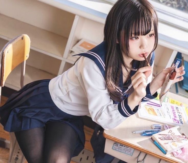 the image shows a young japanese schoolgirl wearing a classic sailor uniform. she's sitting at a desk in what appears to be a classroom. her hair is long and black, styled with bangs. she is eating a chocolate stick and appears to be studying, with textbooks and school supplies around her.  the overall aesthetic is very cute and reminiscent of anime style.  she is looking slightly away from the camera while holding a small mirror.