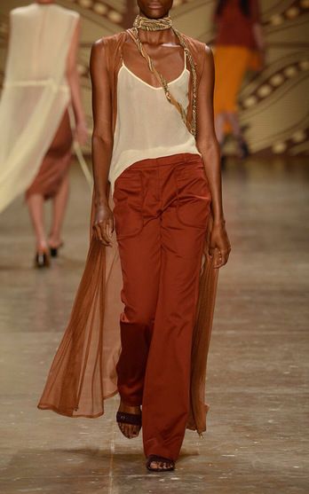 a fashion model walks down the runway in a chic outfit. she is wearing a flowing white top with a sheer long, rustcolored jacket and rustcolored pants. she is accessorized with a long gold chain necklace and black heeled sandals.  the model's hair is pulled back and she is wearing minimal makeup.  the model is walking with confidence and elegance as she showcases the latest fashion trends.