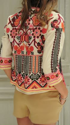 a woman in a white longsleeved embroidered cardigan with red, orange, black, and yellow details is wearing a pair of beige shorts. she is standing outdoors and smiling, giving off a cool and confident vibe. her outfit is stylish and trendy, making her look like a model or a fashion icon. the image has a warm and positive vibe, reflecting the joy and happiness of the woman.