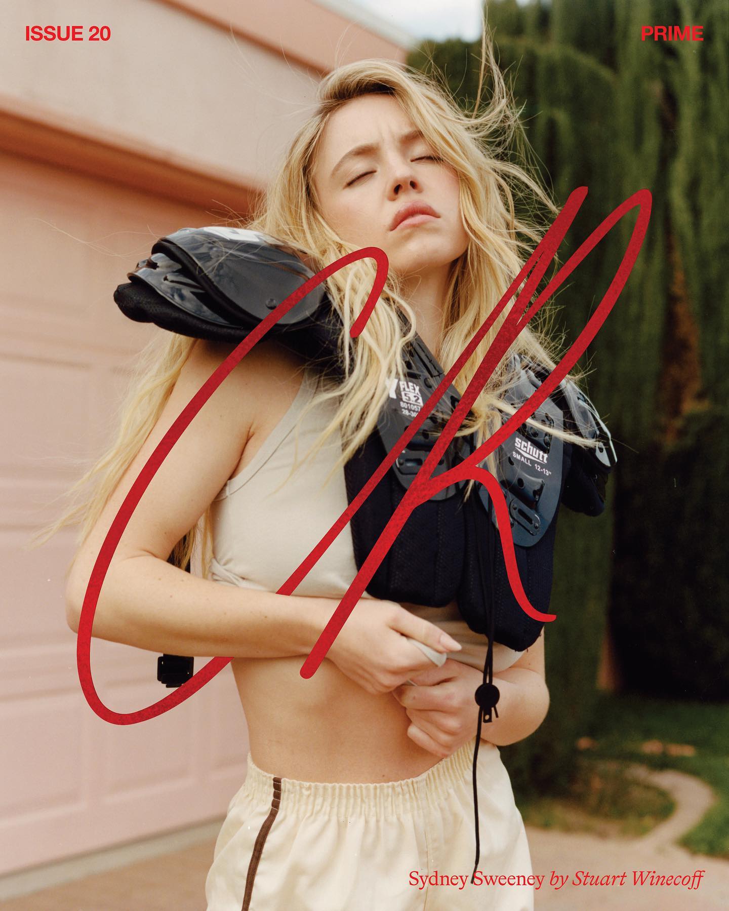 the image is a magazine cover featuring actress sydney sweeney for cr magazine, issue 20.  it's a stylized portrait shot outdoors, with sweeney wearing a beige sports bra and matching shorts.  she has on black football shoulder pads, adding a contrast to her feminine attire. her blonde hair is styled loosely. the background features a pink wall, some greenery, and a hint of a garage. the cr logo is overlaid in red, styled with a dynamic, handwrittenstyle font. the photographer, stuart winecoff, is credited at the bottom. the overall mood is a mix of feminine and athletic, with a glamorous edge.