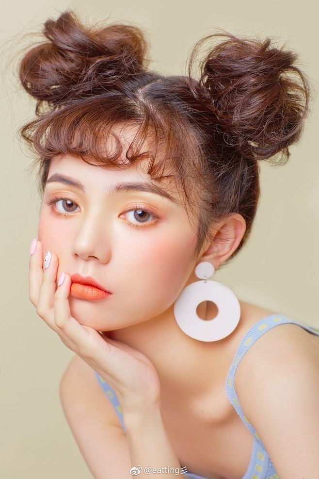 a closeup portrait of a young asian woman with her hair styled in two high space buns. she is wearing minimal pastel makeup featuring blush and orange lipstick. she has large, white, circleshaped earrings. her expression is soft and gentle. she is wearing a light blue polka dot sleeveless top. the background is a light, neutral beige. the overall aesthetic is soft, feminine, and summery.