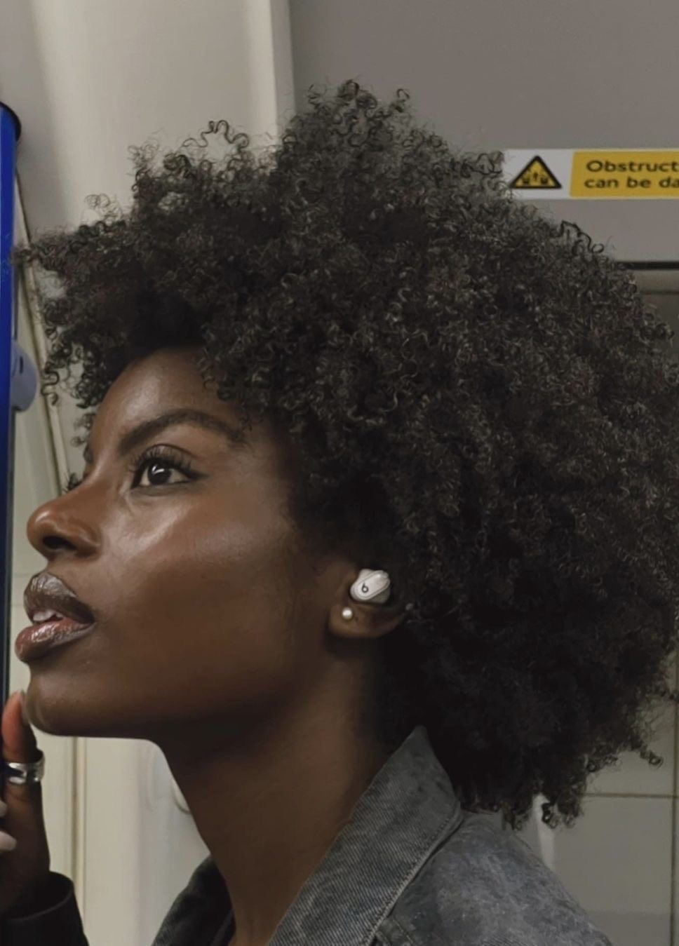 a young black woman with an afro hairstyle, is wearing an earbud and looking down. she is wearing a grey denim jacket. she's inside what looks like a train station.