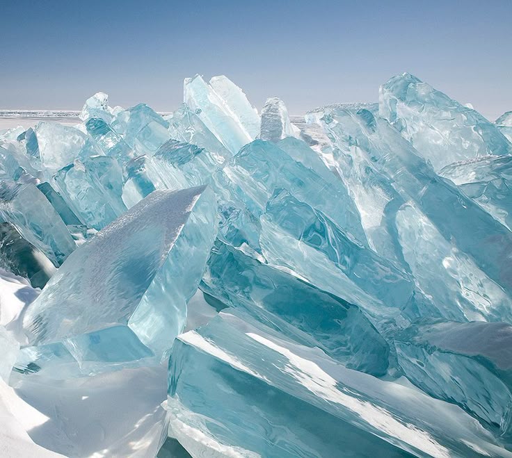 the image shows a pile of large, blue ice blocks on a snowy surface under a clear blue sky. the ice is transparent and has a shardlike appearance. the scene evokes a sense of cold and winter.