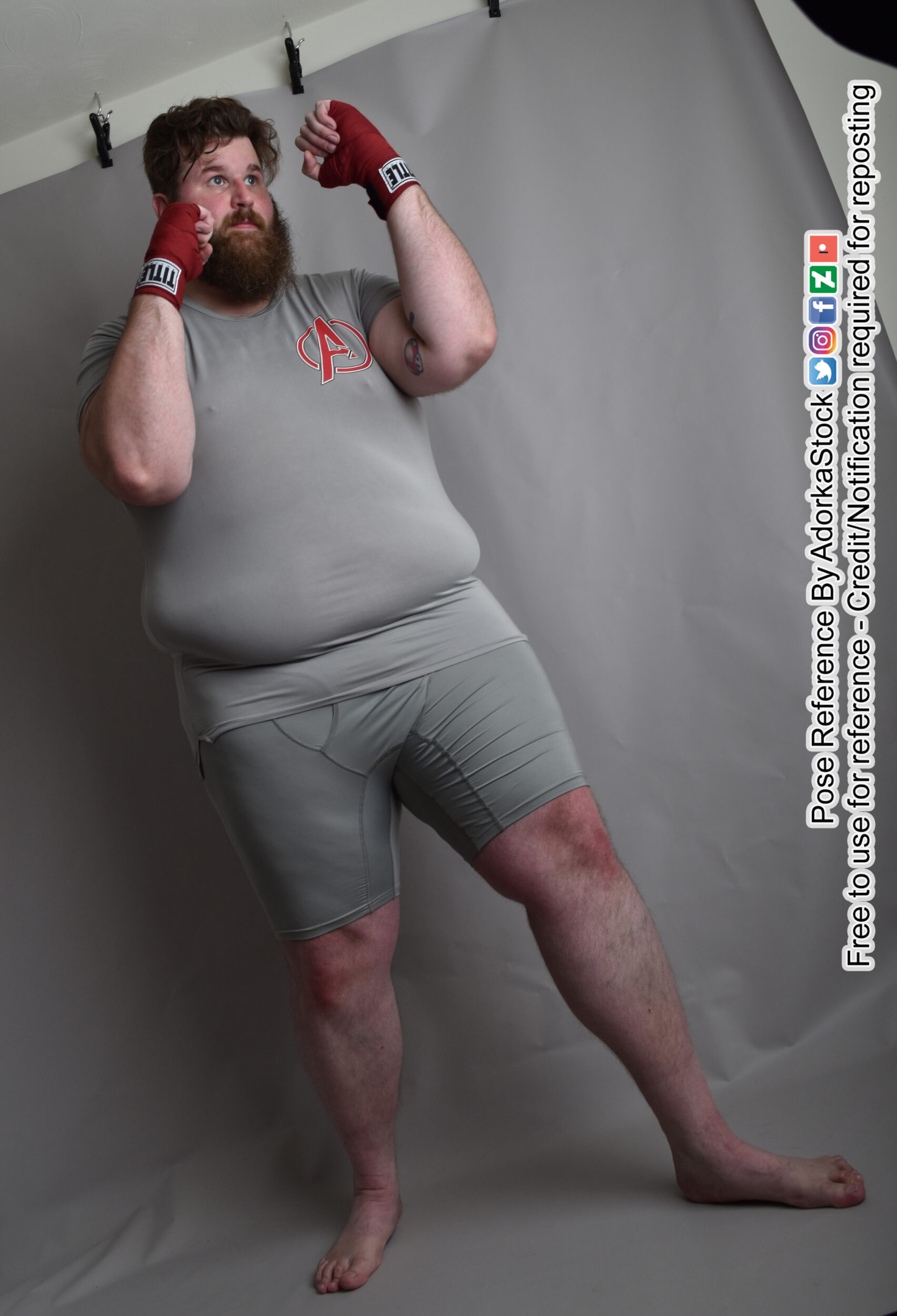 a muscular man in grey shorts and shirt is standing on a grey background.  he is holding his right arm up, wearing a boxing glove,  in a boxing stance with his left arm at his side and his left hand holding a boxing glove as well. he is looking to the right of the camera. this photo could be used for reference for artists and animators.