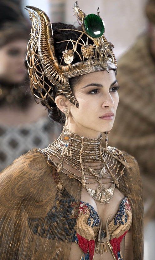 closeup of a woman dressed as an egyptian queen or goddess. she wears an elaborate gold headdress adorned with an emerald gemstone, intricate gold necklaces and a stunning, ornate gold and jewelencrusted bodice. her expression is serious and regal. the background is blurred but shows hints of other people and an ancient setting, suggesting a film still or artistic representation. the overall image exudes richness and power, embodying a sense of ancient grandeur.