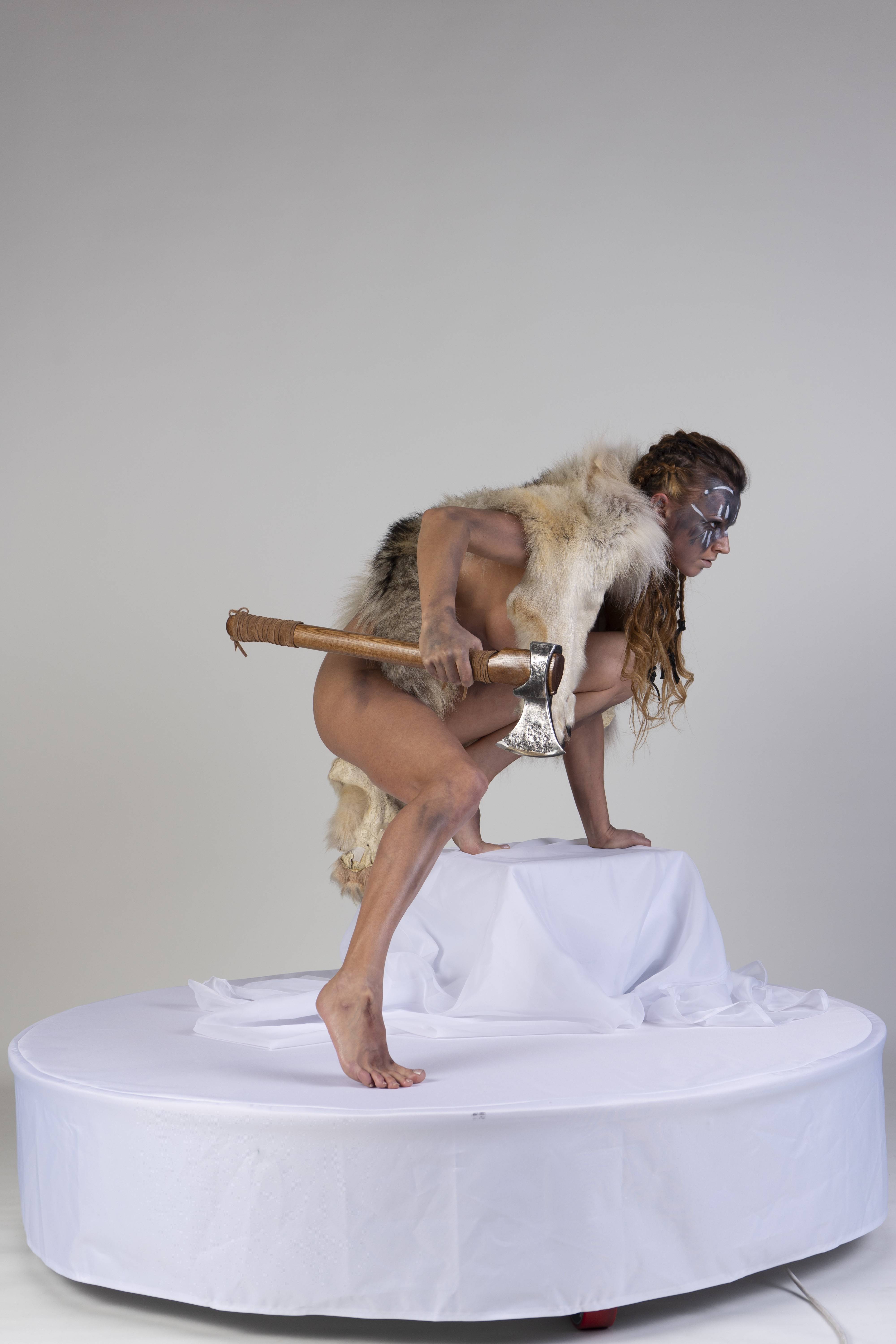 a male warrior, nude except for a fur garment, is crouching on a white platform. he is holding an axe and has face paint. the background is a plain studio backdrop.