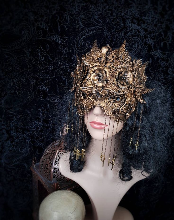 a mannequin head is wearing a golden decorative mask that covers the upper half of its face. the mask is intricately designed with floral patterns, and it has chains hanging down with small crosses attached to them. the mannequin has a dark wig with curly hair. next to the mannequin is a dark wooden decorative item and a skull. the background is a dark patterned material.