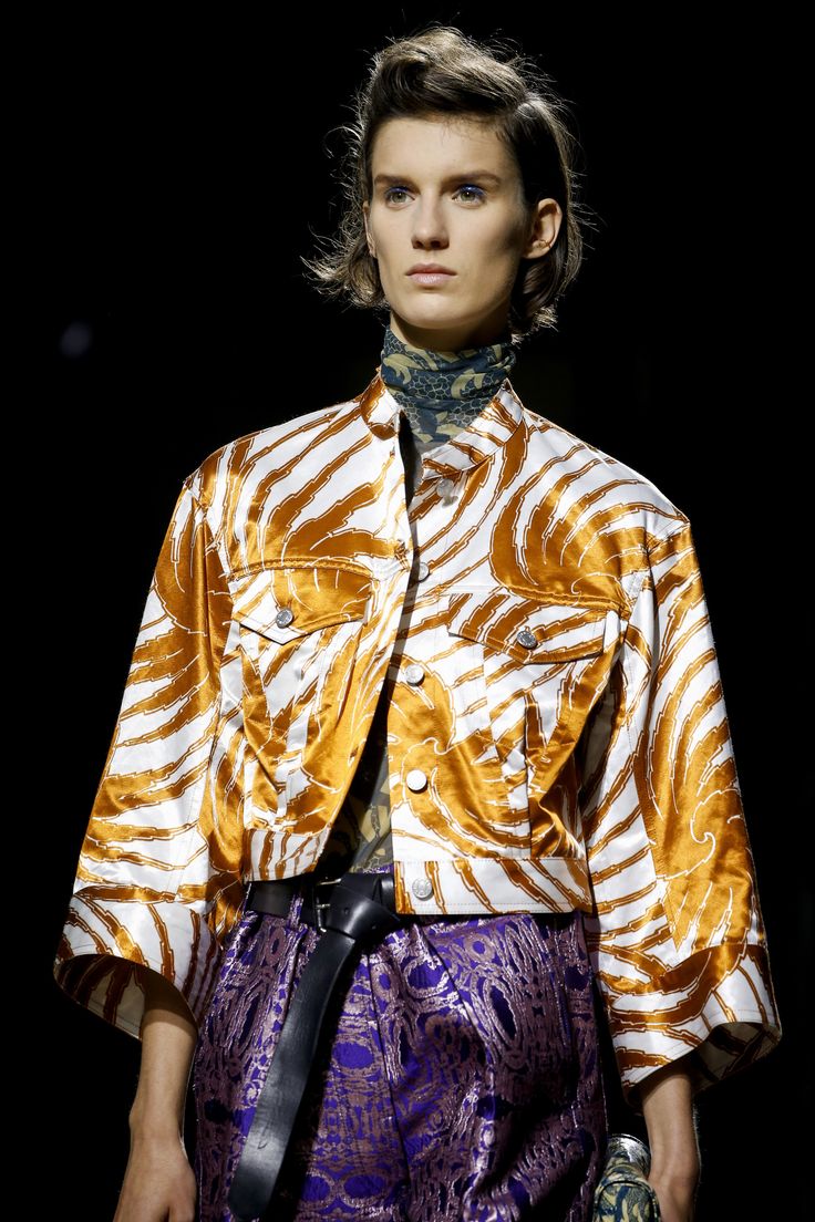 a fashion model with dark hair styled in a voluminous updo showcases a unique ensemble on a runway.  she's wearing a cropped, orange and white satin jacket with a bold, swirling zebralike pattern. underneath, she sports purple brocade pants, cinched with a wide black leather belt.  her turtleneck is a darker, patterned fabric peeking from under the jacket's collar. the background is dark and out of focus, emphasizing the model and her outfit. the overall impression is one of high fashion and a bold, sophisticated style.
