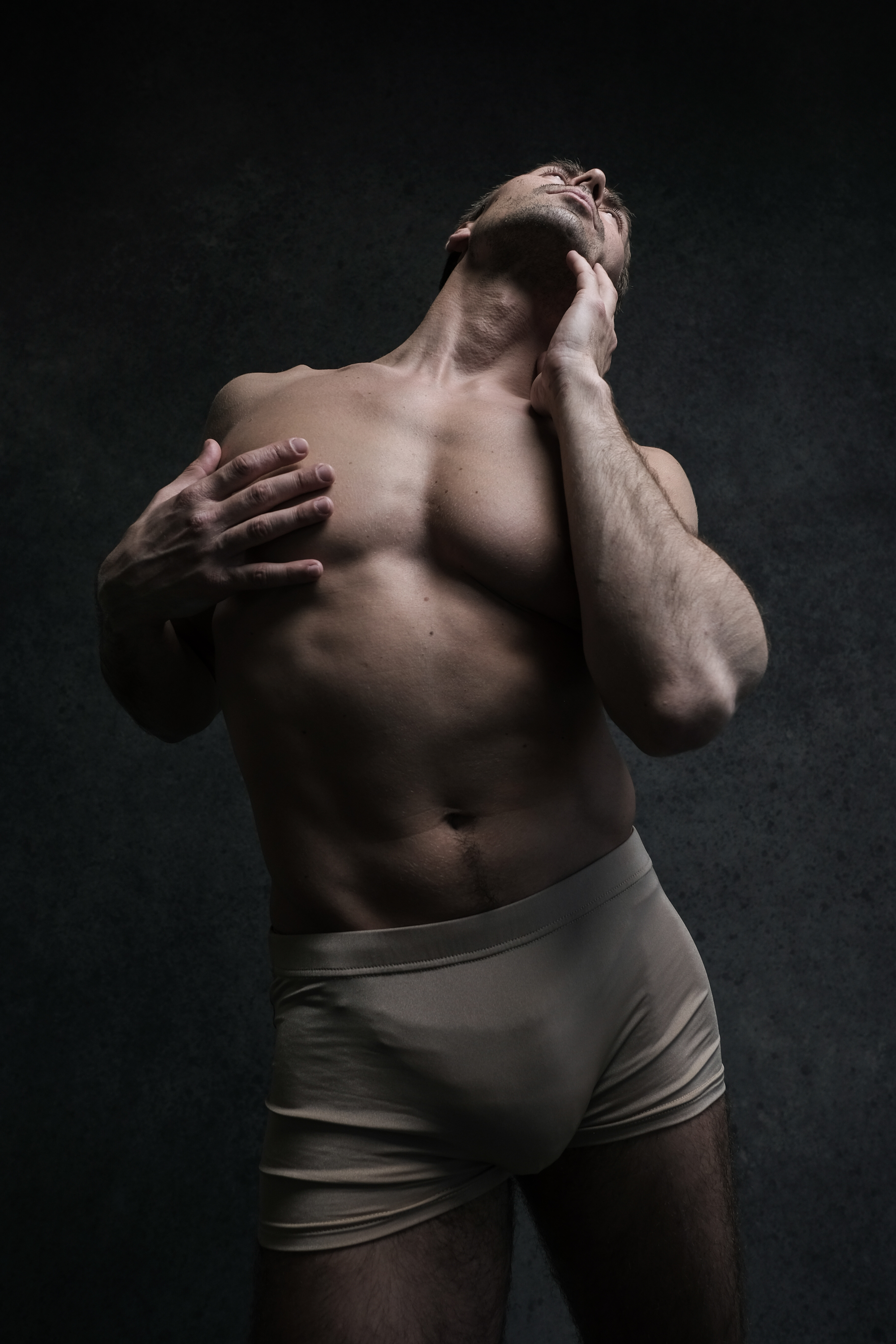 muscular man in beige underwear stands against a dark gray background, his head tilted back and eyes closed. his pose is sensual and expressive, highlighting his physique. the lighting is dramatic, creating strong shadows and emphasizing his muscles. the low camera angle further accentuates his strength and masculinity.
