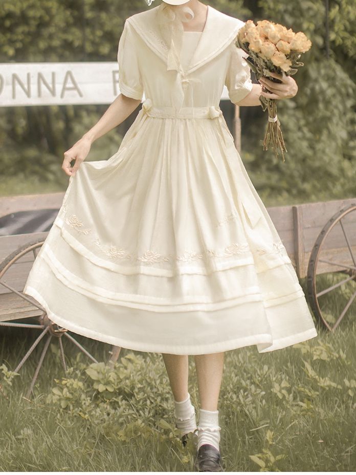 a woman in a vintage style cream dress with a sailor collar is standing in a field. she is holding a bouquet of yellow flowers and her dress is blowing in the wind. there is a wooden wagon visible behind her. the overall aesthetic is soft, romantic, and whimsical.