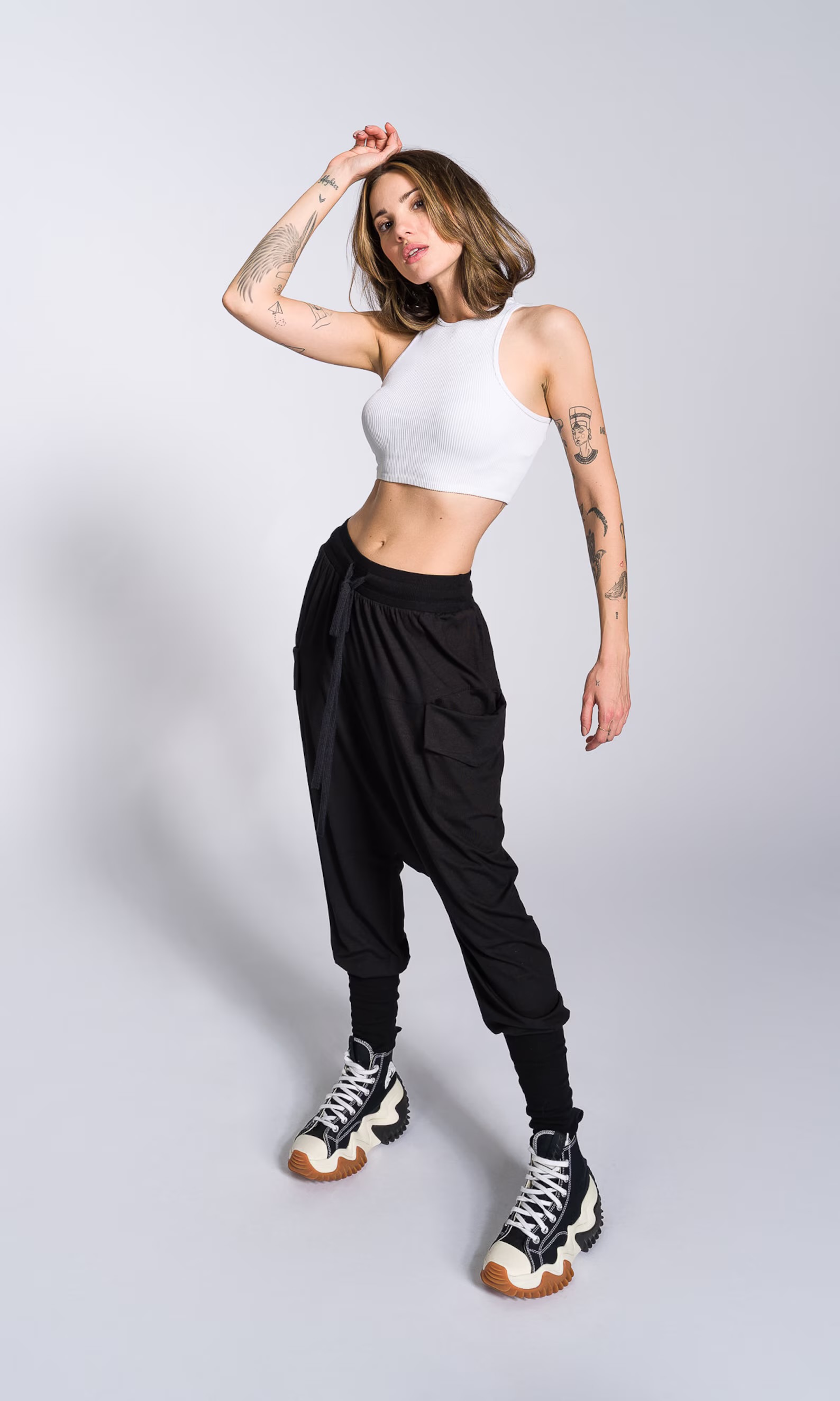 a fullbody shot of a young woman posing in a studio against a light grey background. she is wearing a white crop top, black harem pants, and hightop sneakers. her arms are adorned with tattoos. her pose is relaxed yet confident. the overall style is casual and trendy.