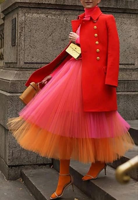a woman is wearing a red coat with gold buttons and a layered tulle skirt in pink and orange. she is carrying a small gold handbag and wearing high heels. she is standing on stairs in an urban setting. she has on a red turtleneck and looks stylish and fashionable.