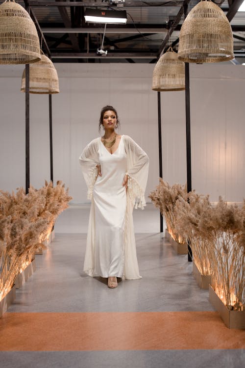 a fashion model is showcasing a stunning bohemianinspired wedding dress on a runway. the dress is long, ivorycolored, features long sleeves with fringe detailing and a sleek slip dress underneath. the model is confidently posing, her dark hair pulled back. the runway is decorated with pampas grass arrangements, creating a neutraltoned and elegant atmosphere.  large, woven pendant lights hang overhead, further enhancing the boho chic style. the overall aesthetic is minimal yet impactful, highlighting the beauty of the dress and model.