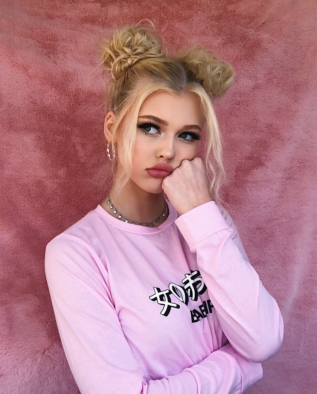 a young blonde woman with her hair styled in space buns poses against a pink backdrop. she's wearing a pink longsleeved shirt with black japanese text, a silver necklace, and matching earrings. her makeup is prominent, and she has a slightly bored or pouting expression with her hand resting on her chin, looking off to the side.