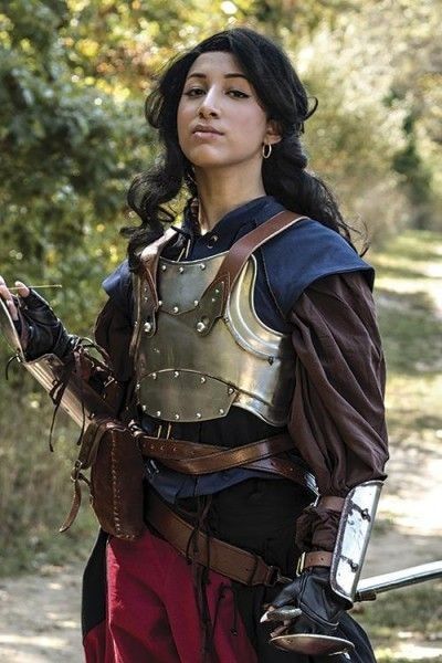 a woman in medievalstyle armor stands outdoors, holding a sword. she is wearing a breastplate over a blue and brown tunic, with a red skirt and a leather belt with pouches. she has black hair and is looking forward. the background is a blurred forest.