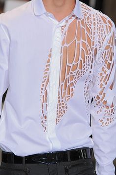 a man is wearing a white shirt with a cutout design that resembles a wing pattern. he is also wearing a black belt with his pants. he is standing in a formal setting, perhaps on a runway or during a photoshoot. his shirt is designed to be stylish and eyecatching. the pattern on his shirt is a key feature of the design.