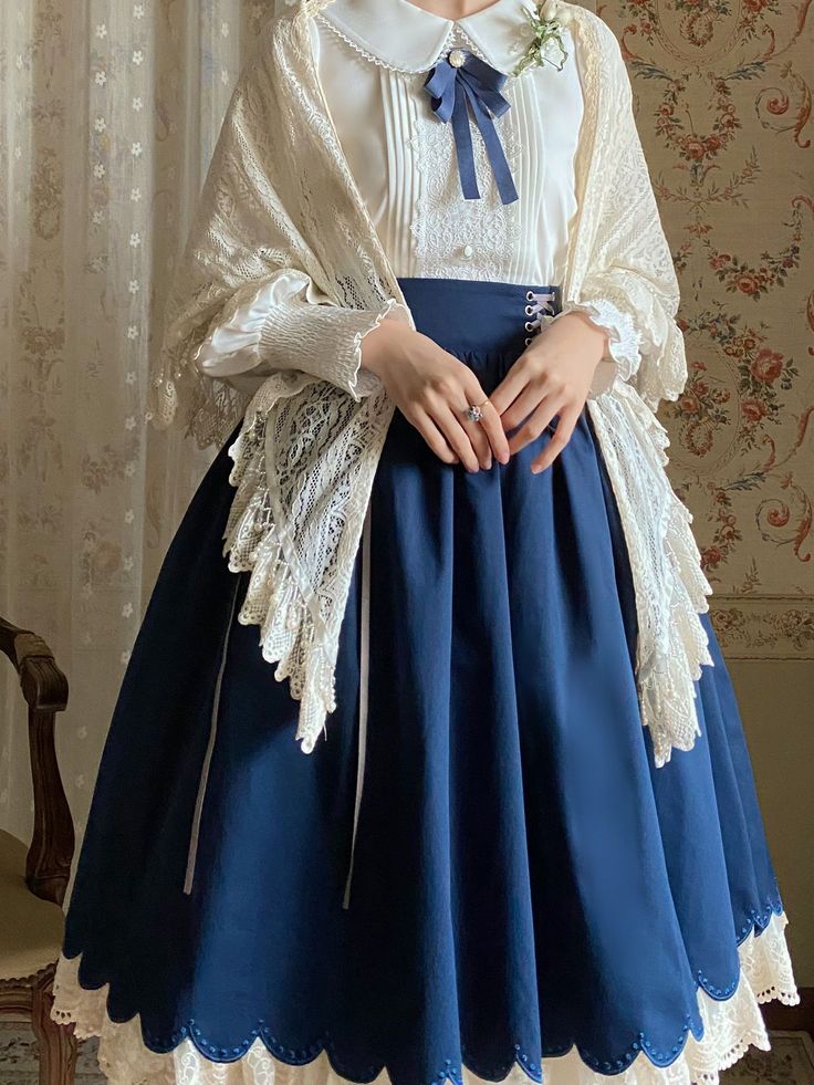 a woman is standing in a room, wearing a navy blue lolita skirt with white lace trim, a white blouse with a peter pan collar, and a cream lace shawl.  she is also wearing a flower corsage on her left shoulder. the skirt is long and flowy, and the blouse has long sleeves. the shawl is draped over her shoulders and hangs down to her waist. the woman is posing with her hands clasped in front of her. the room is decorated in a vintage style, with floral wallpaper and a wooden chair. the lighting is soft and natural.