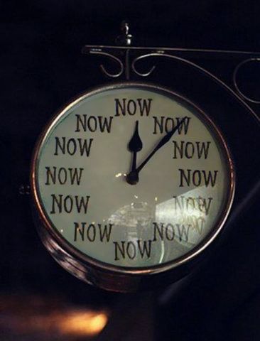a vintage clock with the word "now" repeated twelve times in place of the numbers. the clock has a black minute and hour hand and is mounted on a wrought iron style bracket.