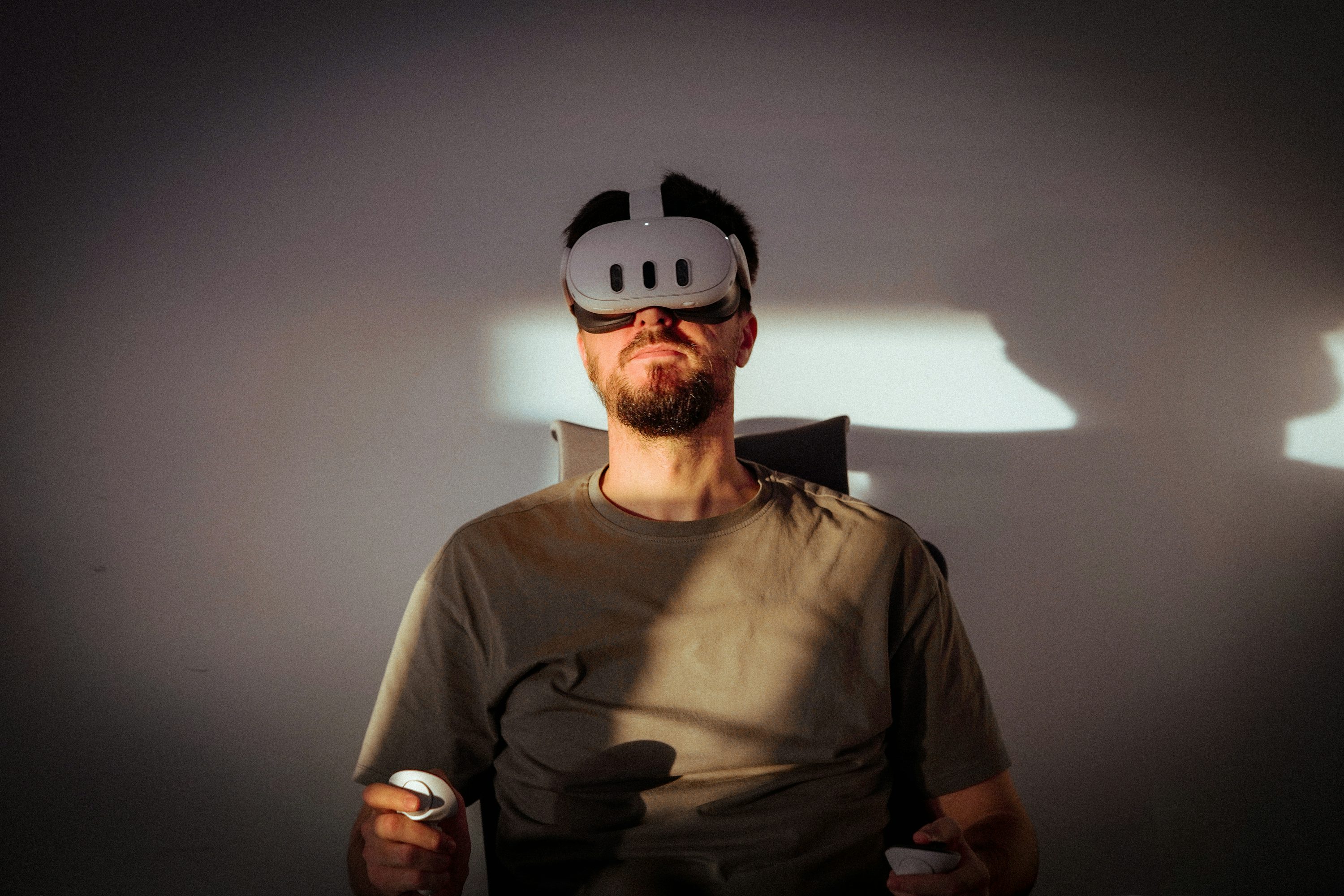 a man wearing a white vr headset is sitting in a chair. he is holding white vr controllers in his hands. the scene is lit with strong shadows cast on the wall behind him. he is wearing a tshirt.