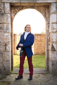 a fullbody shot of a man standing in a stone archway. he is wearing a blue coat, maroon pants, a white shirt, and black shoes. he is holding a single red rose. the background is a stone ruin, and the lighting is bright and natural. the man's expression is serious and contemplative.