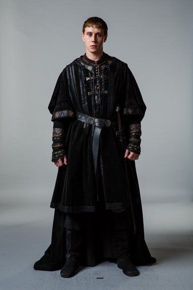 a man stands in a studio wearing medieval clothing. he is wearing a long, black tunic with a leather belt and a sword. he is standing with his hands at his sides and looking directly at the camera. the background is a solid gray color.