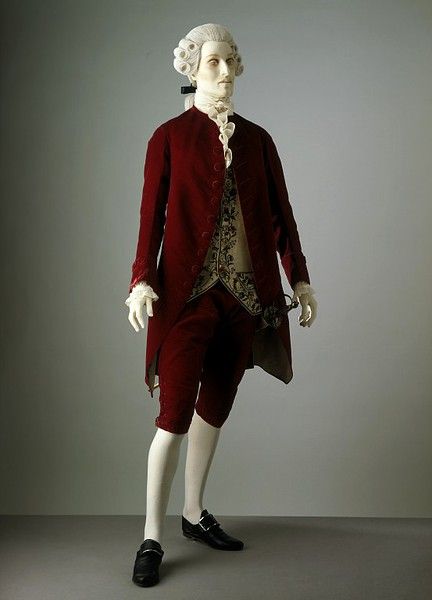 a mannequin dressed in 18thcentury men's fashion. the mannequin wears a white wig with curls, a white jabot at the neck, a red suit consisting of a long jacket and breeches, and a decorative waistcoat. the mannequin also wears white stockings and black buckled shoes. the background is a plain gray.