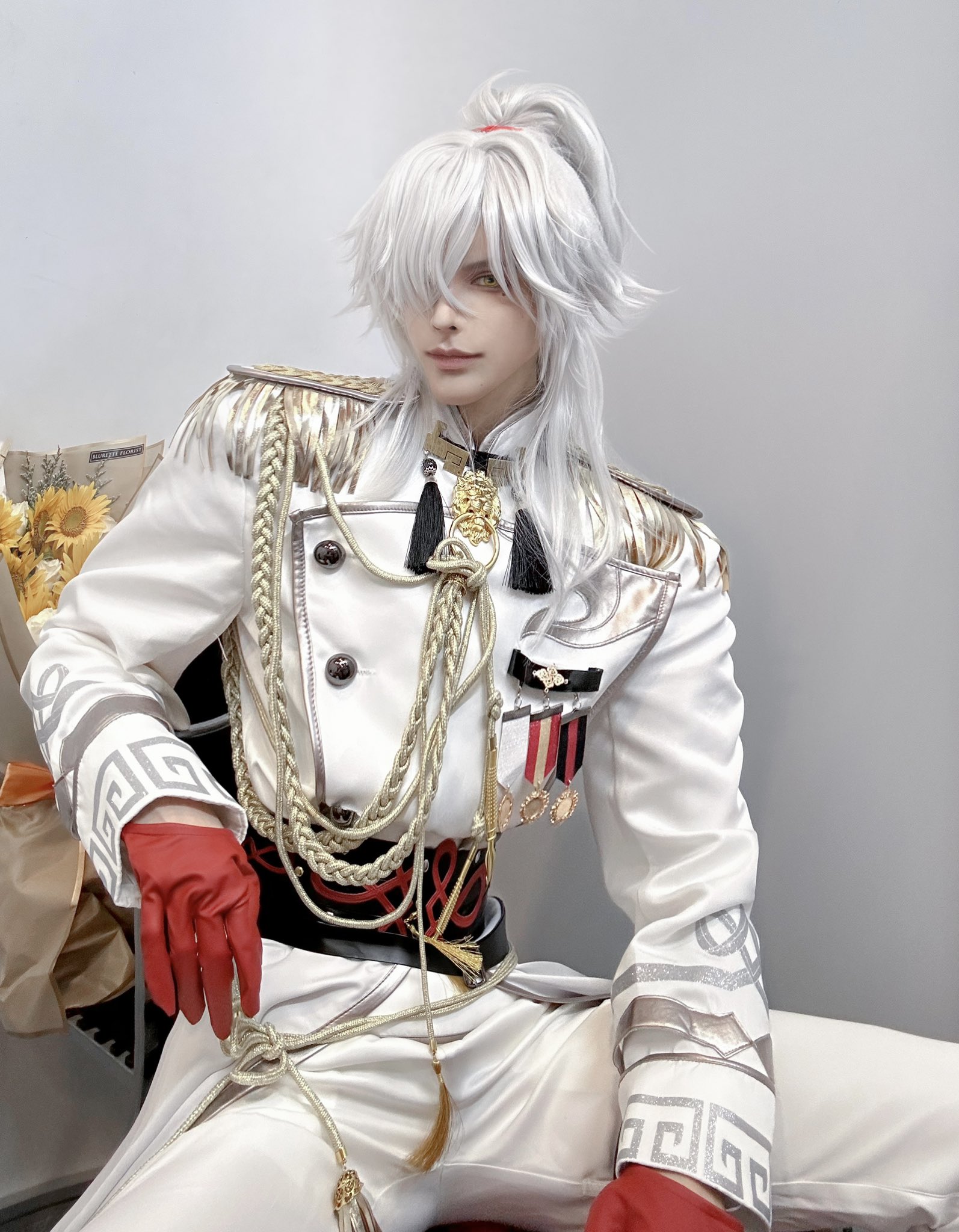 a young man cosplaying an anime character with white hair, wearing a white suit with gold accents and red gloves. he has a serious and elegant expression. he is sitting in a chair with his legs crossed. the background is blurry and white.