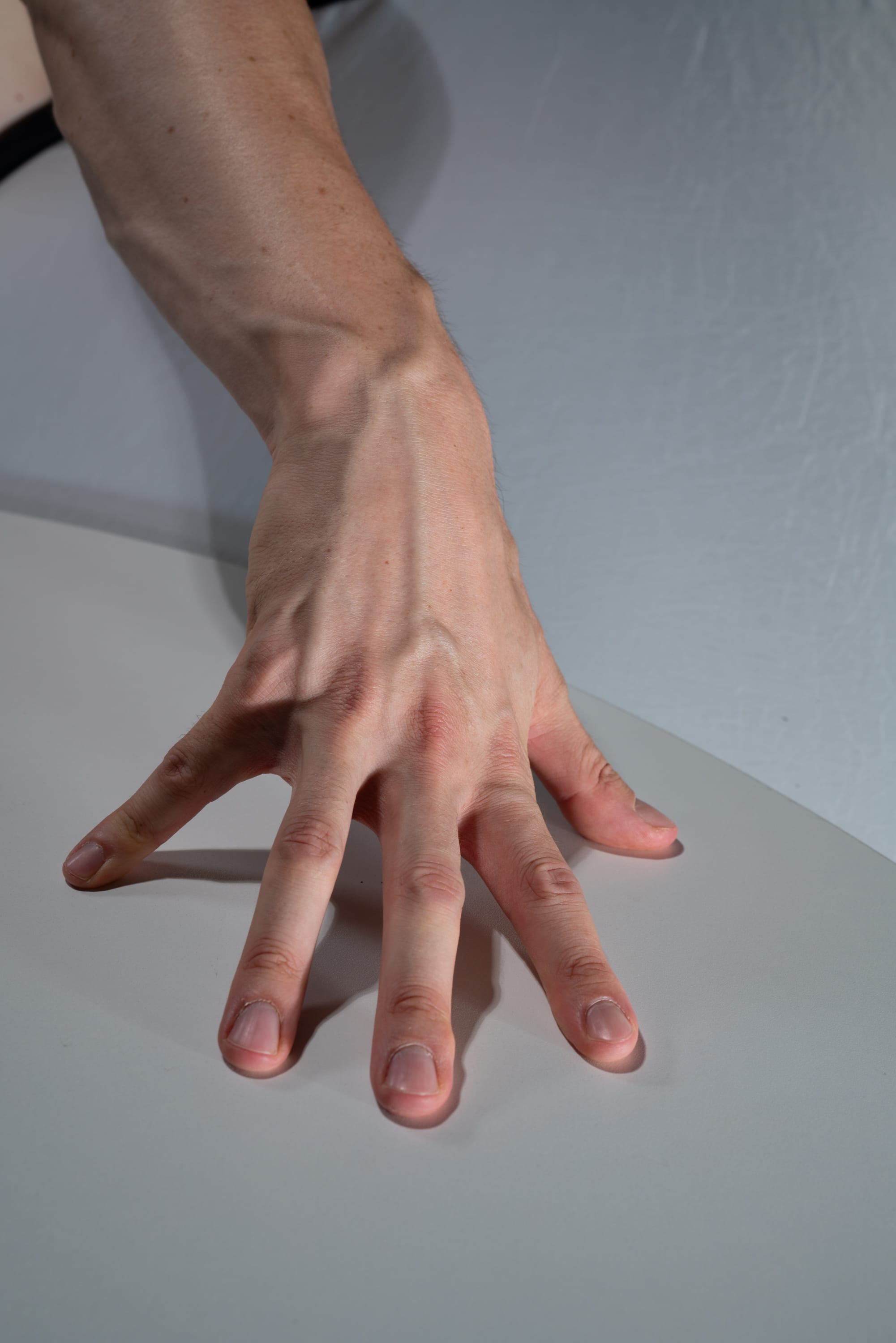 a caucasian male hand is outstretched on a white table, fingers splayed.  the studio lighting emphasizes the contours of the hand, highlighting prominent veins and tendons.