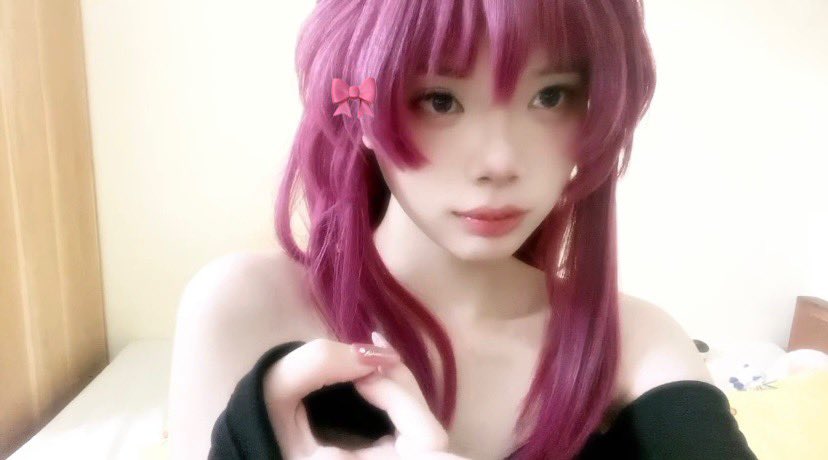 a young woman with long pink hair, wearing black clothing, is sitting in a bedroom with a white bed. she has a red bow in her hair and is looking at the camera.