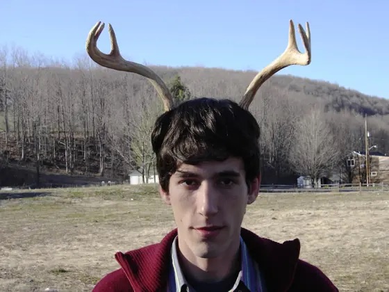 a young man is wearing a pair of antlers as a headband. he is smiling and looking at the camera. he is outdoors in a field with trees and mountains in the background.