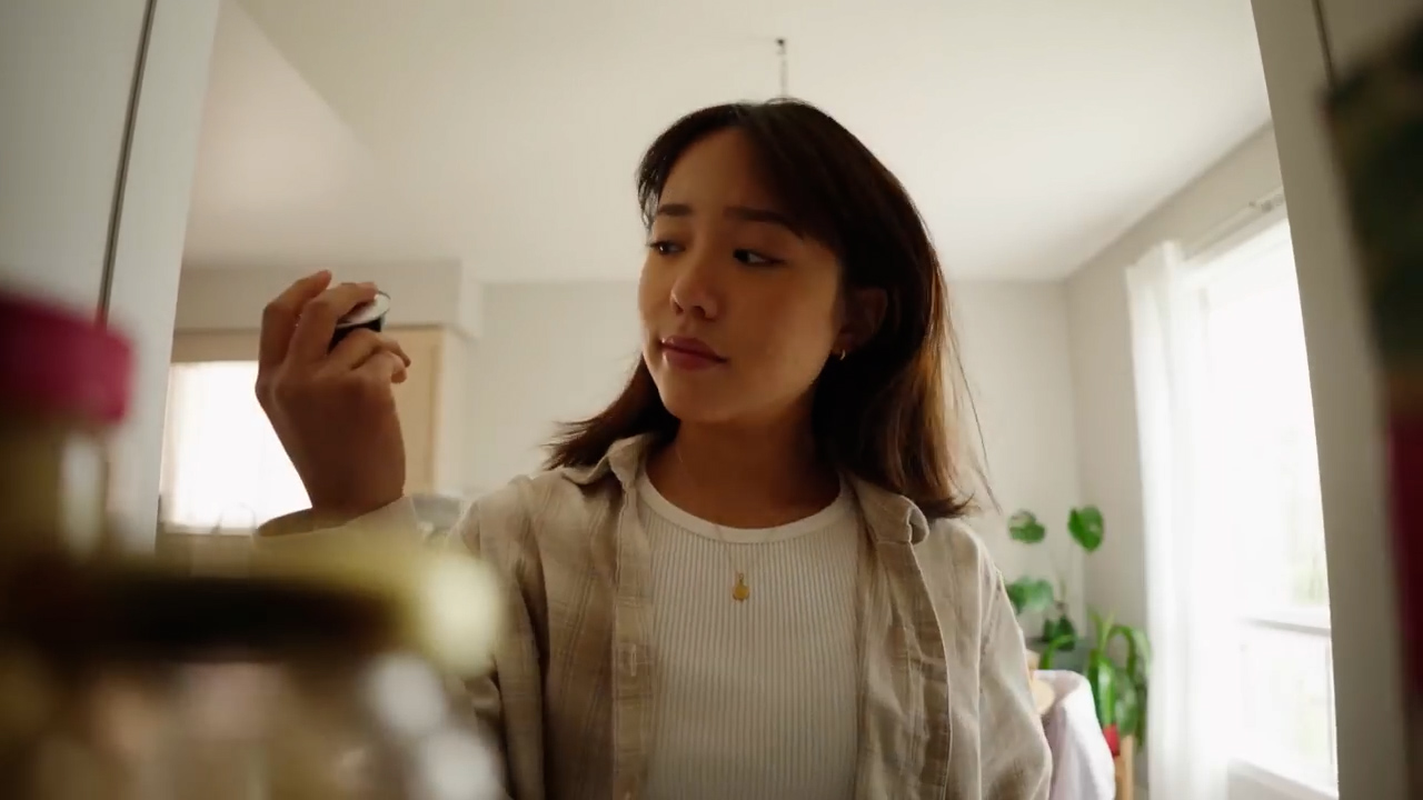 a young woman with shoulderlength black hair is pictured from the perspective of someone inside a refrigerator. she's wearing a white tshirt and a light brown overshirt. she is looking at something in her hand and her expression is neutral. the room she's standing in is welllit with natural light and has white walls. there is a green plant visible to the right of the frame.